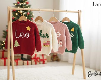 May include: Four long-sleeved knit sweaters in red, cream, pink, and green hanging on a wooden rack. Each sweater features embroidered names and festive patches including a star, train, snowman, and bell. The text Lamoriea Embroidery is visible.