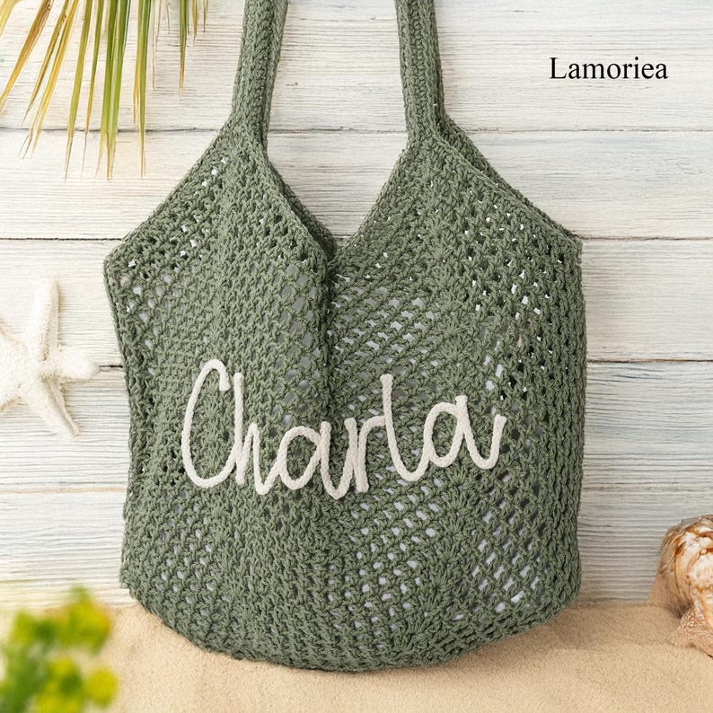 Peut inclure: Un sac fourre-tout en crochet vert avec le nom "Charla" en &eacute;criture blanche. Le sac a deux poign&eacute;es et une large ouverture. Le mot "Lamoriea" est imprim&eacute; dans le coin sup&eacute;rieur droit. Le sac est pos&eacute; sur du sable.