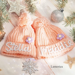 Op de afbeelding: Twee perzikkleurige gebreide mutsen met pompons, elk voorzien van een kleine lavendelkleurige bloemapplicatie. De linker muts is geborduurd met de naam Stella in wit schrift, en de rechter muts is geborduurd met de naam Maddie in wit schrift.