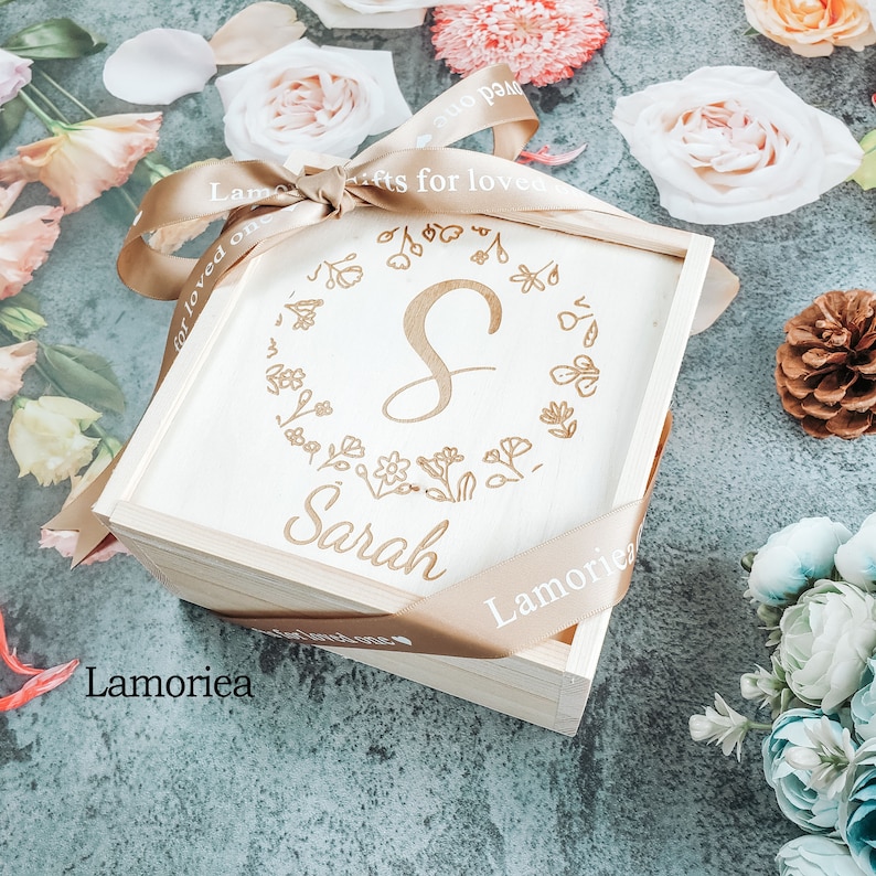 May include: A wooden box with a light brown ribbon tied around it. The box is engraved with the letter "S" surrounded by a floral design and the name "Sarah". The ribbon is labeled "Gifts for loved ones" and "Lamoriea".