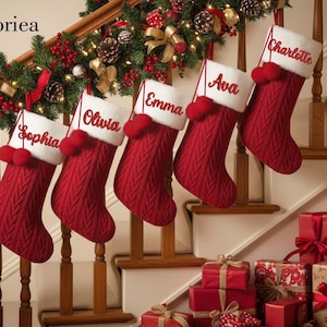 May include: Five red knitted Christmas stockings with white cuffs and personalised names hang on a staircase. Each stocking has a red pom-pom. A garland with red berries and pinecones decorates the banister. Wrapped gifts are at the base of the stairs.