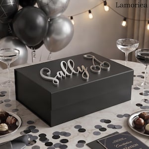 May include: A rectangular black gift box featuring the name Sally in silver script lettering and a silver cherry icon on the lid. The box is positioned on a table with scattered black confetti, two clear glasses, and a small plate of chocolates.