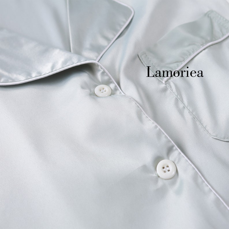 Puede incluir: Primer plano de una camisa de pijama de sat&eacute;n azul claro con ribetes y botones blancos. La camisa tiene un bolsillo en el pecho y el nombre de la marca "Lamoriea" impreso.