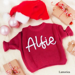 May include: A deep red long-sleeve knit jumper featuring the name Alfie embroidered in white cursive script across the chest. The jumper has a ribbed crew neckline and ribbed cuffs, displayed alongside a red Santa hat and gift boxes.