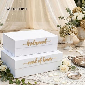 Könnte beinhalten: Zwei weiße Geschenkboxen, eine aufeinander gestapelt. Auf der oberen Box steht "bridesmaid" in goldener Schrift, und auf der unteren Box steht "maid of honor". Die Boxen stehen auf einer weißen Oberfläche mit Blumenarrangements.
