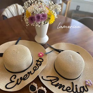Könnte beinhalten: Zwei beige Stroh-Sonnenhüte mit schwarzen Bändern und den Namen "Everest" und "Amelia" in Schreibschrift. Jeder Hut hat eine kleine dekorative Blume. Eine weiße Vase mit bunten Blumen und Sonnenbrillen sind ebenfalls vorhanden.