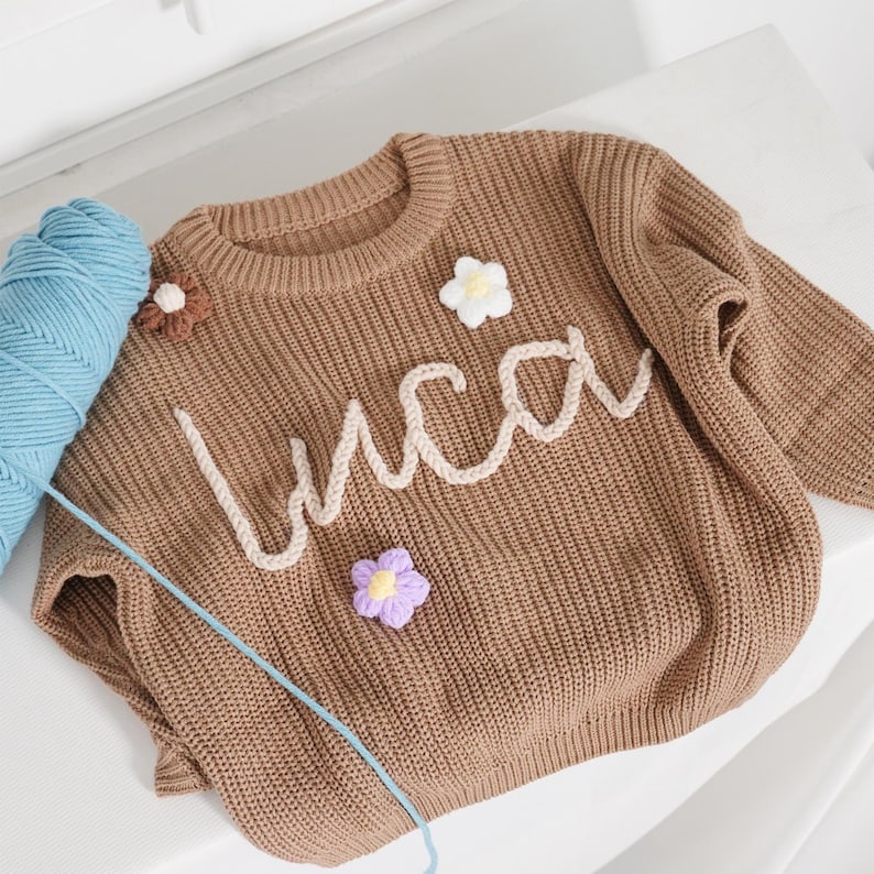 May include: A brown long-sleeve knit jumper featuring the name "Luca" in cream-coloured yarn lettering across the chest. The jumper is decorated with three small, crocheted flower appliqu&eacute;s in white, brown, and purple, and sits next to a ball of blue yarn.