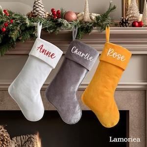Puede incluir: Tres calcetines de Navidad personalizados con nombres bordados en el puño. Los calcetines son blancos, grises y amarillos.
