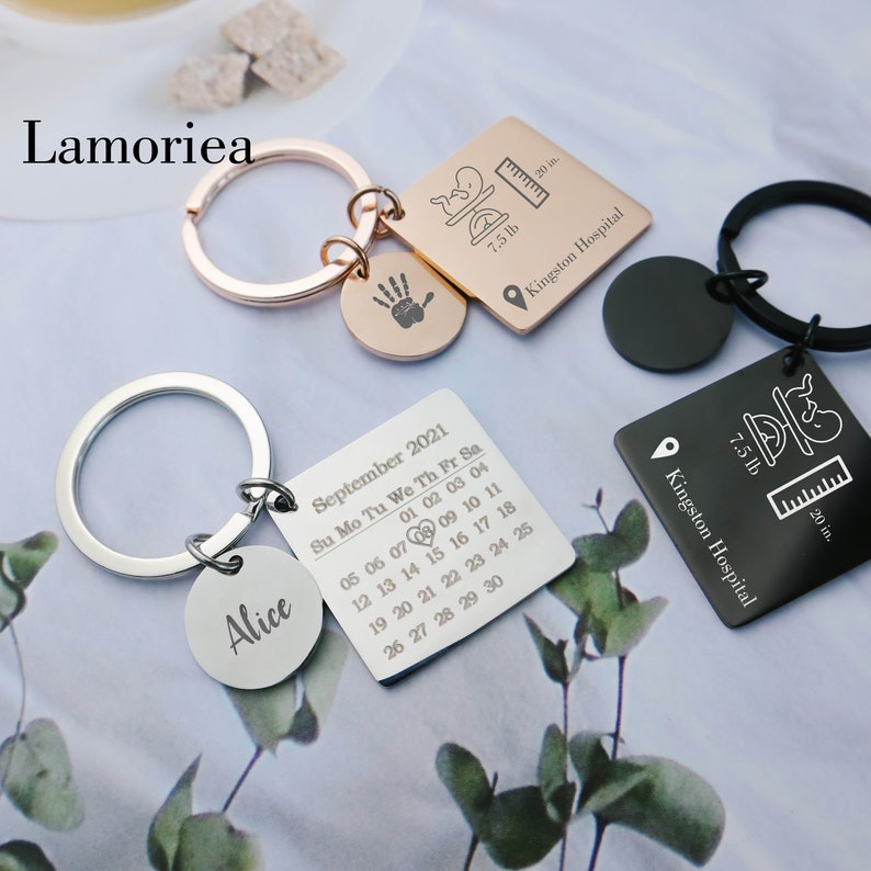 May include: Two silver keyrings with engraved details. One keyring features a calendar with the month of September 2021 and the name "Alice". The other keyring has a baby footprint, a ruler, and the text "7.5 lb" and "Kingston Hospital".