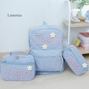 May include: A light blue corduroy backpack, lunch bag, and pencil case set. Each item features the name "Josie" in pink script and white flower accents. The brand name "Lamoriea" is in the upper left corner.