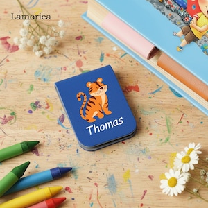 Puede incluir: Un cuaderno azul personalizado con una ilustraci&oacute;n de un tigre de dibujos animados y el nombre "Thomas" en blanco. El cuaderno est&aacute; sobre una superficie de madera con crayones y un libro en el fondo. Es un art&iacute;culo para ni&ntilde;os.