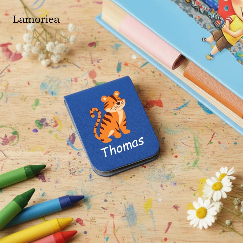 Puede incluir: Un cuaderno azul personalizado con una ilustraci&oacute;n de un tigre de dibujos animados y el nombre "Thomas" en blanco. El cuaderno est&aacute; sobre una superficie de madera con crayones y un libro en el fondo. Es un art&iacute;culo para ni&ntilde;os.