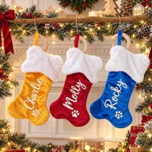 Pode incluir: Três meias de Natal em forma de osso em tecido de veludo amarelo, vermelho e azul. Cada meia apresenta um punho de pele sintética branca, um nome bordado em branco e uma pata branca. Os nomes são Charlie, Molly e Rocky.