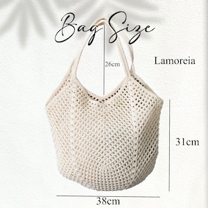 Peut inclure: Sac fourre-tout en crochet blanc cass&eacute; avec une ouverture en forme de V. Le sac mesure 38 cm de large, 31 cm de haut et les anses mesurent 26 cm de long. Le texte "Bag Size" et "Lamoreia" sont visibles. Le sac a un design ouvert tiss&eacute;.