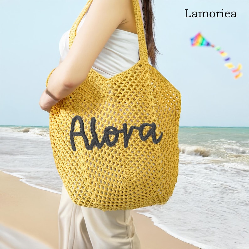 Peut inclure: Un sac fourre-tout jaune en crochet avec le mot "Alora" en lettres gris fonc&eacute;. Le sac a un grand motif ajour&eacute; et de longues bretelles. L'arri&egrave;re-plan montre une sc&egrave;ne de plage avec un cerf-volant.
