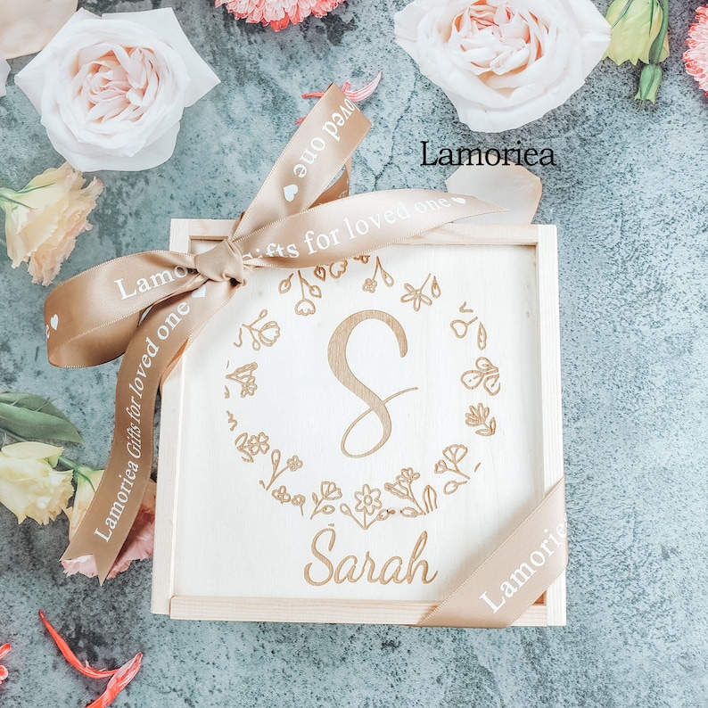May include: A wooden box with a light brown ribbon tied around it. The box is engraved with the letter "S" surrounded by a floral design and the name "Sarah". The ribbon has the text "Lamoriea Gifts for loved one" printed on it.