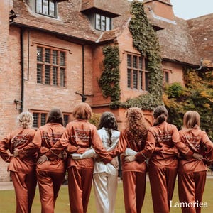 May include: A group of seven people wearing matching long-sleeved button-down pajama sets in rust orange and white. Each shirt features white embroidered text on the back, including names and dates, with a heart icon and floral accents.
