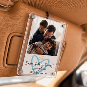 Könnte beinhalten: Ein klarer Acryl-Autosonnenblenden-Fotorahmen mit einem Familienfoto. Das Foto zeigt einen Vater, der zwei Kinder umarmt. Der Text "Drive Safely Daddy! Love you xx Alex & Andrew" ist auf dem Foto aufgedruckt.