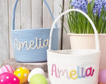 May include: Two white rope Easter baskets with the name Amelia embroidered on them in different colored yarn. The basket on the left is light blue and the basket on the right is white.