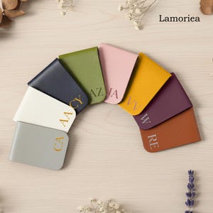 Puede incluir: Una colección de carteras de cuero personalizadas en varios colores, incluyendo gris, blanco, azul marino, verde, rosa, amarillo, morado y marrón. Cada cartera está grabada con iniciales en letras doradas o plateadas. La marca "Lamoriea" es visible.