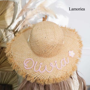 Peut inclure: Un chapeau de soleil en paille naturelle avec un large bord et des bords effilochés. Le chapeau porte le nom "Olivia" brodé en rose en écriture cursive, ainsi qu'une petite fleur rose. La marque "Lamoriea" est visible.