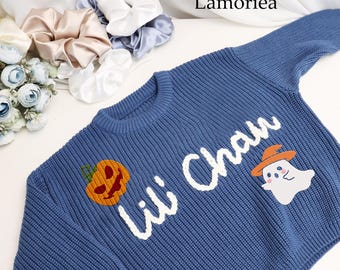 May include: A blue knitted sweater with a white embroidered design that says "Lili Chau". The sweater has a pumpkin and a ghost with a hat embroidered on it.