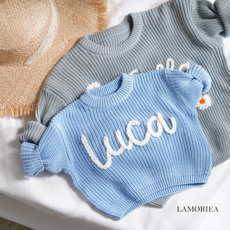 May include: A light blue knit jumper featuring the name Luca in white cursive yarn lettering, layered over a grey knit jumper with a white daisy flower detail, accompanied by a tan straw hat with a frayed edge and a white ribbon trim.
