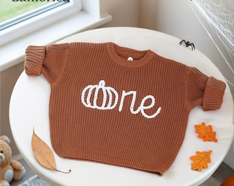 May include: A brown knit sweater with the word "one" and a pumpkin design in white. The sweater is on a white table with decorative leaves and spider decorations. The brand name "Lamoriea" is in the top left corner.