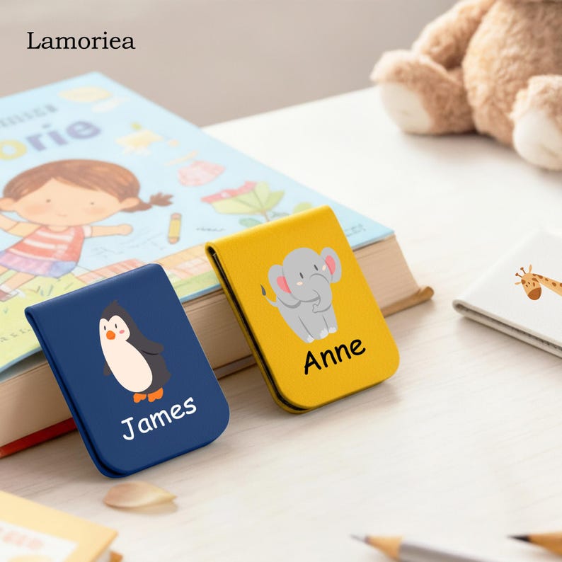 Puede incluir: Dos cubiertas de libros personalizadas, una azul marino con un ping&uuml;ino y el nombre "James", y otra amarilla con un elefante y el nombre "Anne". Un libro infantil est&aacute; en el fondo.