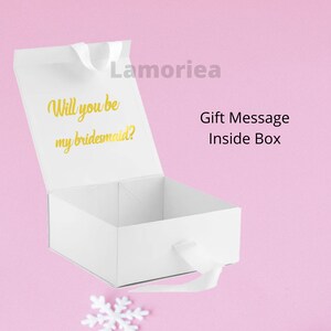 Może przedstawiać: Białe pudełko prezentowe z złotą wstążką i tekstem "Will you be my bridesmaid?" wewnątrz pokrywy. Pudełko jest otwarte, a pokrywa jest uniesiona. Tekst "Gift Message Inside Box" jest nadrukowany na zewnątrz pudełka.