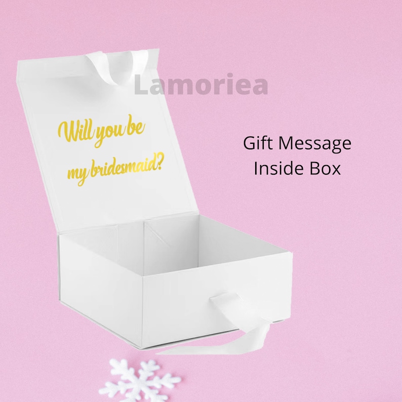 Może przedstawiać: Białe pudełko prezentowe z złotą wstążką i tekstem "Will you be my bridesmaid?" wewnątrz pokrywy. Pudełko jest otwarte, a pokrywa jest uniesiona. Tekst "Gift Message Inside Box" jest nadrukowany na zewnątrz pudełka.