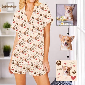 Op de afbeelding: Een tweedelige pyjamaset bestaande uit een overhemd met korte mouwen en knoopsluiting en een bijpassende short. De crèmekleurige stof heeft een herhalend patroon van bruine hondengezichten, pootafdrukken, rode hartjes en de naam Buddy.