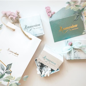 May include: A set of four gift boxes with a marble pattern and gold lettering that says "Lamoreia Jewelry". The boxes are on a white surface with green leaves and pink flowers.