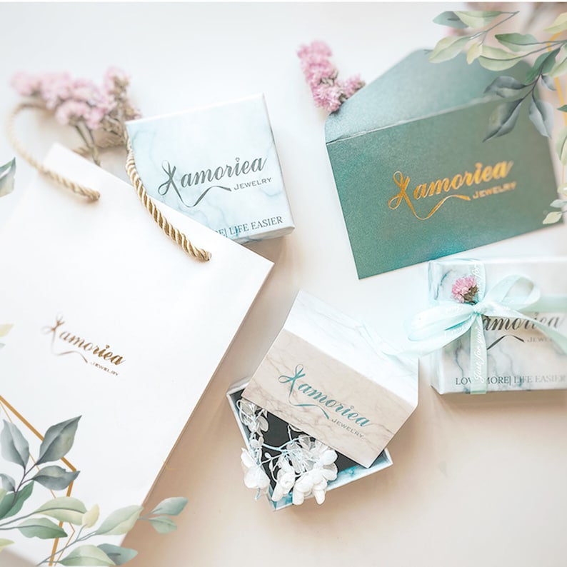 May include: A set of four gift boxes with a marble pattern and gold lettering that says "Lamoreia Jewelry". The boxes are on a white surface with green leaves and pink flowers.