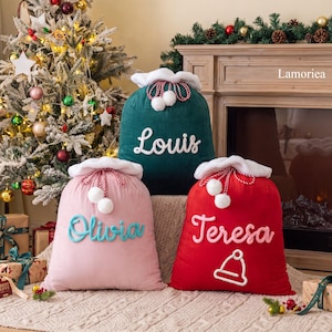 Pode incluir: Três sacos de presente de Natal de veludo com acabamento em pele sintética branca e cordões trançados vermelhos e brancos com pompons. O saco verde tem o nome Louis, o rosa o nome Olivia e o vermelho o nome Teresa.