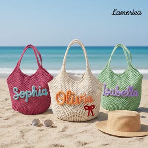 May include: Three crochet tote bags in pink, cream, and green, each with a name embroidered on the front: Sophia, Olivia, and Isabella. A straw hat and sunglasses sit on the sand. The bags are on a beach with the ocean in the background.