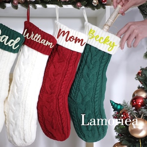 May include: Four cable-knit Christmas stockings in white, red, and green, each with a white cuff and a name embroidered in a contrasting color: "Dad", "William", "Mom", and "Becky". The stockings are hanging from a decorative branch.