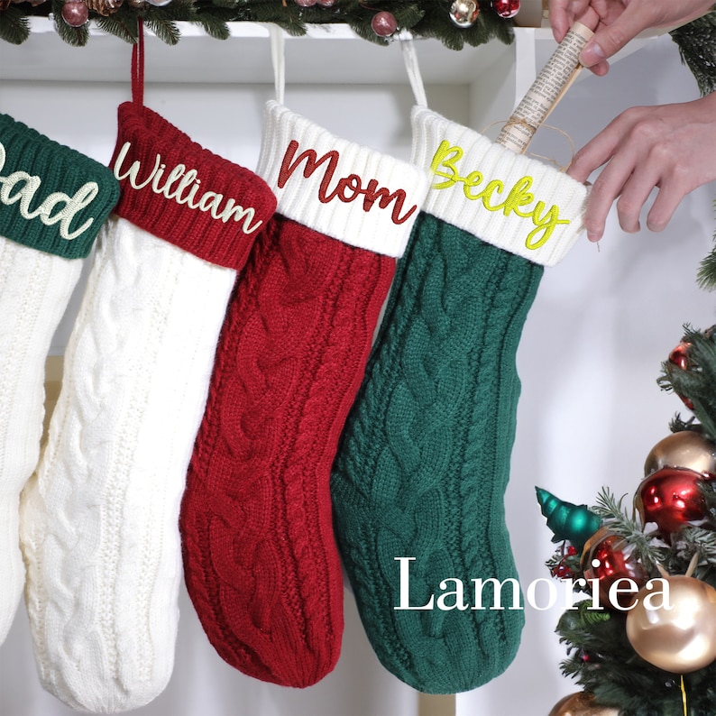 May include: Four cable-knit Christmas stockings in white, red, and green, each with a white cuff and a name embroidered in a contrasting color: "Dad", "William", "Mom", and "Becky". The stockings are hanging from a decorative branch.