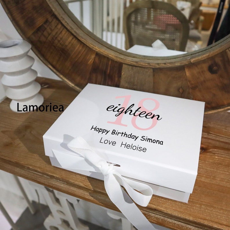 May include: White gift box with a pink and black "eighteen" design. The box has a white ribbon and the text "Happy Birthday Simona Love Heloise" is printed on the box.