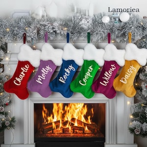 Puede incluir: Una colección de calcetines navideños personalizados en varios colores, incluyendo rojo, morado, azul, verde, magenta y dorado. Cada calcetín tiene un puño blanco y está bordado con un nombre en escritura blanca. Los calcetines cuelgan sobre una chimenea.
