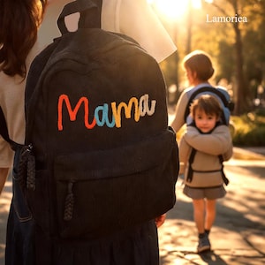 以下が含まれることがあります： カラフルな糸で「ママ」と刺繍された黒いコーデュロイのバックパック。バックパックには前面にジッパー付きポケットと調節可能なストラップが付いています。