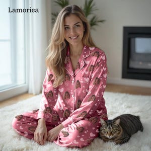 Könnte beinhalten: Rosa Pyjama-Set mit einem sich wiederholenden Muster aus Gesichtern, Kronen und Herzen. Das langärmlige Oberteil hat weiße Paspeln und Knöpfe. Die passende Hose vervollständigt das Set. Der Markenname "Lamoriea" befindet sich oben links.