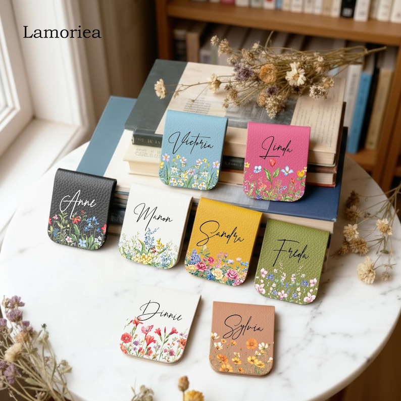 May include: A collection of personalized leather magnetic bookmarks in various colors, each adorned with a floral design and a name in elegant script. The bookmarks are displayed on a marble surface near a stack of books.