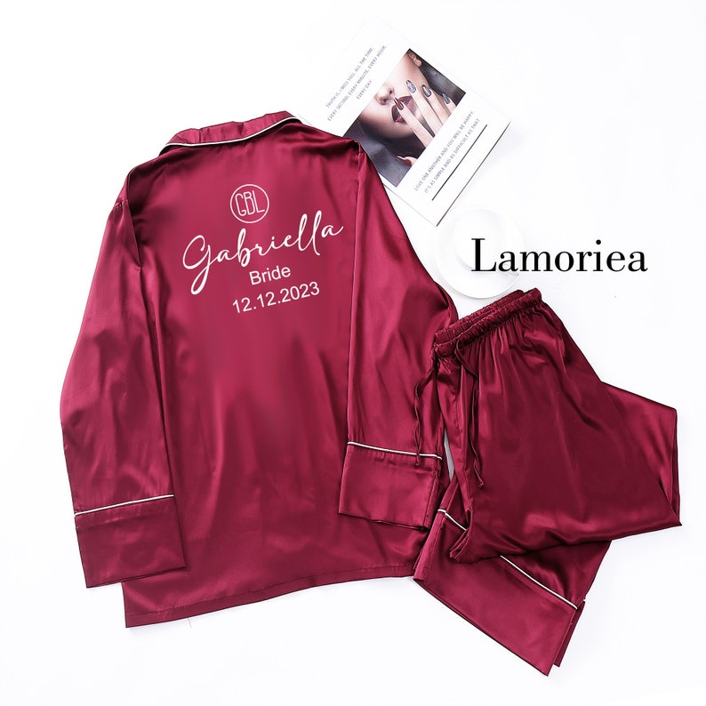 Puede incluir: Conjunto de pijama de sat&eacute;n burdeos con ribete blanco. La parte superior presenta el nombre "Gabriella", la palabra "Bride" y la fecha "12.12.2023" en escritura blanca. La marca "Lamoriea" es visible.