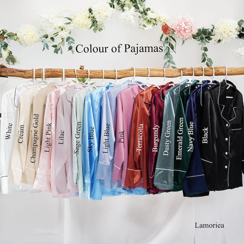 May include: A collection of long-sleeved button-down pajama shirts hanging on a wooden branch, labeled with colors: White, Cream, Champagne Gold, Light Pink, Lilac, Sage Green, Sky Blue, Light Blue, Pink, Terracotta, Burgundy, Dusty Green, Emerald Green, Navy Blue, and Black.