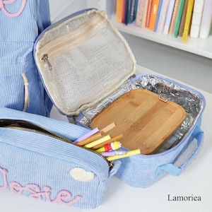 Pu&ograve; includere: Un portapranzo in velluto a coste azzurro chiaro con astuccio e zaino abbinati. Il portapranzo &egrave; aperto, rivelando un tagliere in legno e una tasca a rete. L'astuccio contiene matite e penne colorate. La parola "Josie" &egrave; ricamata sull'astuccio.