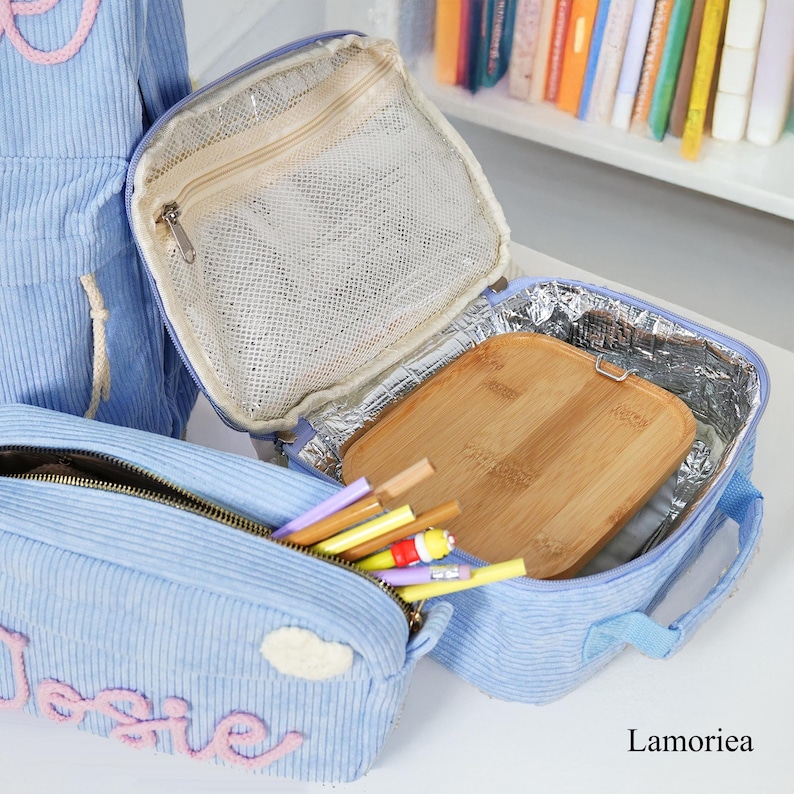 Pu&ograve; includere: Un portapranzo in velluto a coste azzurro chiaro con astuccio e zaino abbinati. Il portapranzo &egrave; aperto, rivelando un tagliere in legno e una tasca a rete. L'astuccio contiene matite e penne colorate. La parola "Josie" &egrave; ricamata sull'astuccio.