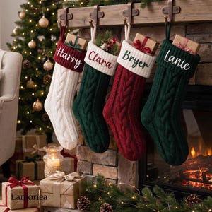 Könnte beinhalten: Vier Weihnachtssocken aus Zopfstrick hängen an einem hölzernen Kaminsims. Die Socken sind weiß, dunkelgrün und tiefrot mit weißen Bündchen, die mit den Namen Harry, Clara, Bryer und Lanie bestickt sind. Jede Socke enthält ein kleines Geschenk.
