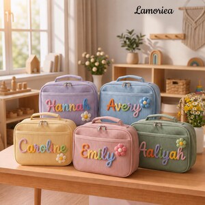May include: Five rectangular corduroy lunch bags in pastel colors: lavender, light blue, yellow, pink, and sage green. Each bag features a top handle, a zippered front pocket, and a name in raised, colorful letters with a small flower applique.