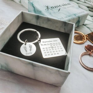 May include: Silver keyring with a round charm featuring a footprint design and the name "Emily" engraved on it. The keyring also includes a square charm with a calendar for September 2021.